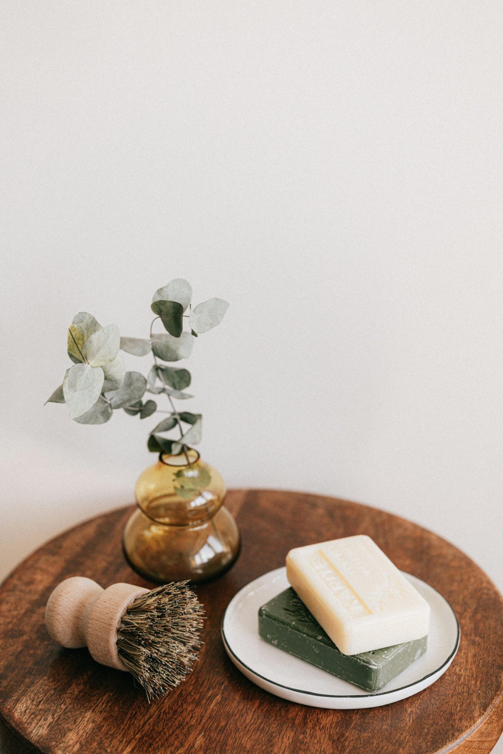 From above composition with assorted organic soaps on white ceramic holder and shaving brush with wooden handle near yellow transparent vase with plant on wooden round table against white wall