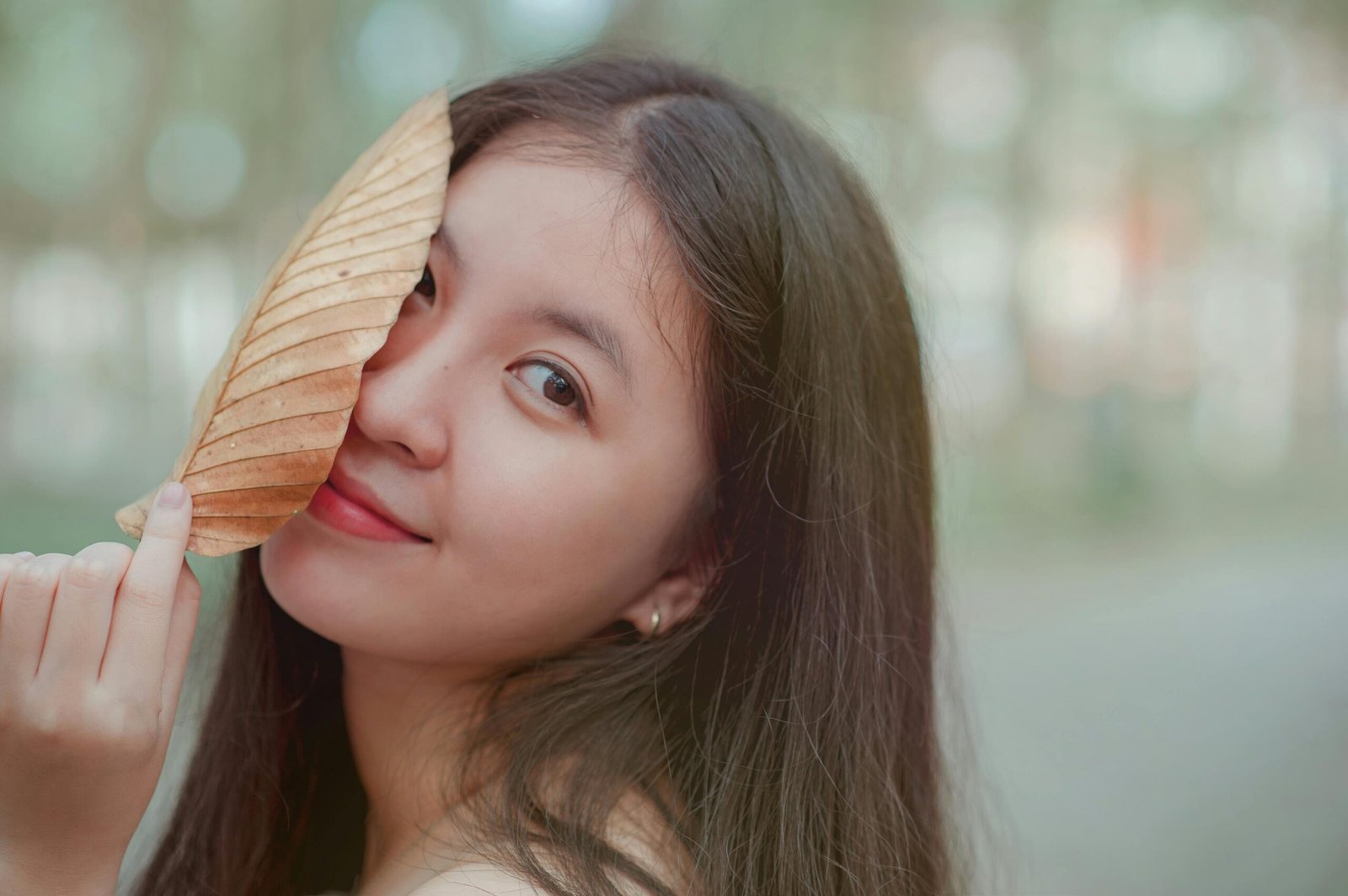 A smiling young woman playfully poses with a dry leaf in a natural outdoor setting.